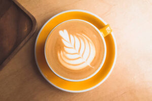 A latte in a yellow cup with a leaf design created in the foam on top.