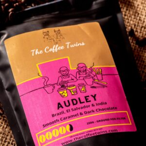 The Coffee Twins Audley coffee bag with a pink label, surrounded by roasted coffee beans. A strong speciality coffee blend with smooth caramel and dark chocolate notes.