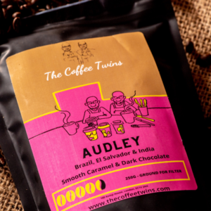 The Coffee Twins Audley coffee bag with a pink label, surrounded by roasted coffee beans. A strong speciality coffee blend with smooth caramel and dark chocolate notes.