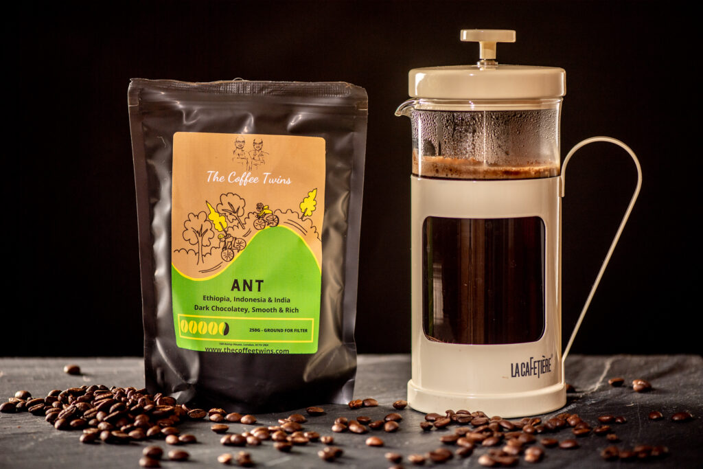 The Coffee Twins bag sat on a black kitchen surface with coffee beans spread out, sat beside a French Press pot of coffee, filled with delicious Ant coffee