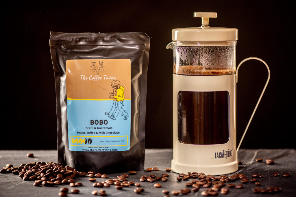 The Coffee Twins bag sat on a black kitchen surface with coffee beans spread out, sat beside a French Press pot of coffee, filled with delicious Bobo  coffee