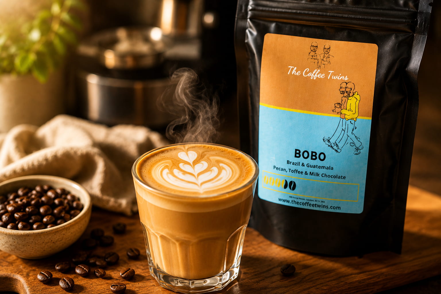 Close-up lifestyle photograph of a freshly made latte with heart-shaped latte art in a clear glass, sitting on a rustic wooden table beside a bag of The Coffee Twins Bobo coffee beans. The scene includes scattered roasted coffee beans, a bowl of coffee beans, and soft steam rising from the coffee, creating a warm, café-style atmosphere that showcases how to make great coffee at home. Ideal image for a home coffee brewing guide and speciality coffee blog.