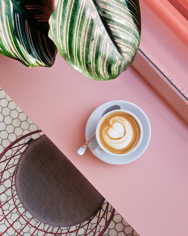 A cup of coffee with latte art on a pink table, accompanied by a spoon.
