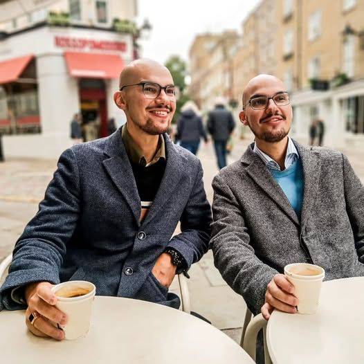 Two men with glasses and bald heads sitting at a table with coffee cups in their hands.