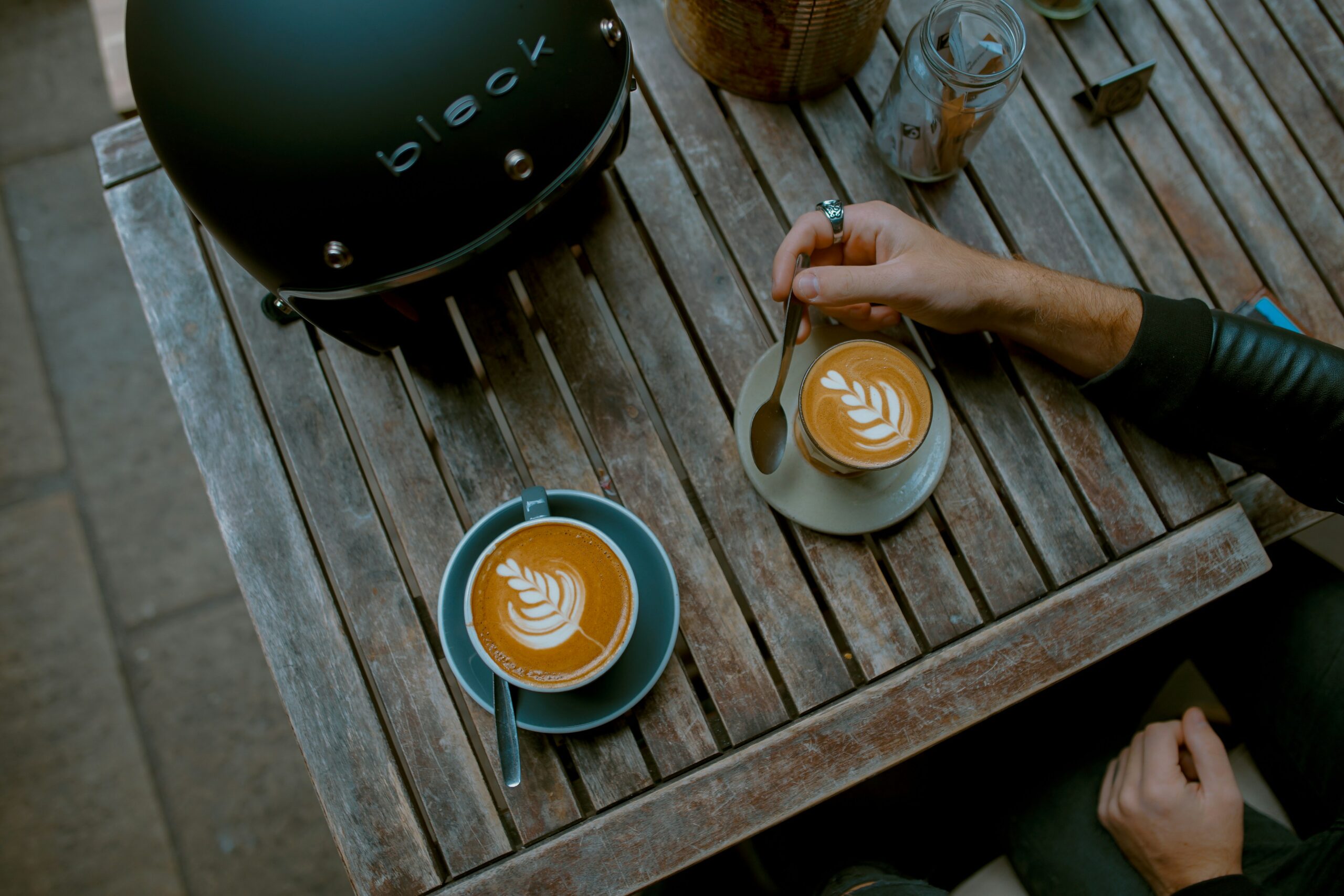 Two cups of coffee with latte art on a wooden table, one cup in a blue saucer and a spoon beside it.