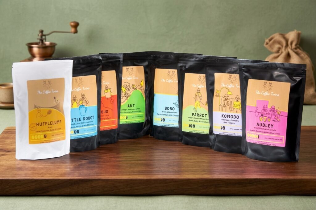 A product lineup of eight colorful "The Coffee Twins" coffee flavor bags, featuring one white bag on the left and seven black bags, displayed on a wooden countertop against a soft green background with a vintage coffee grinder.