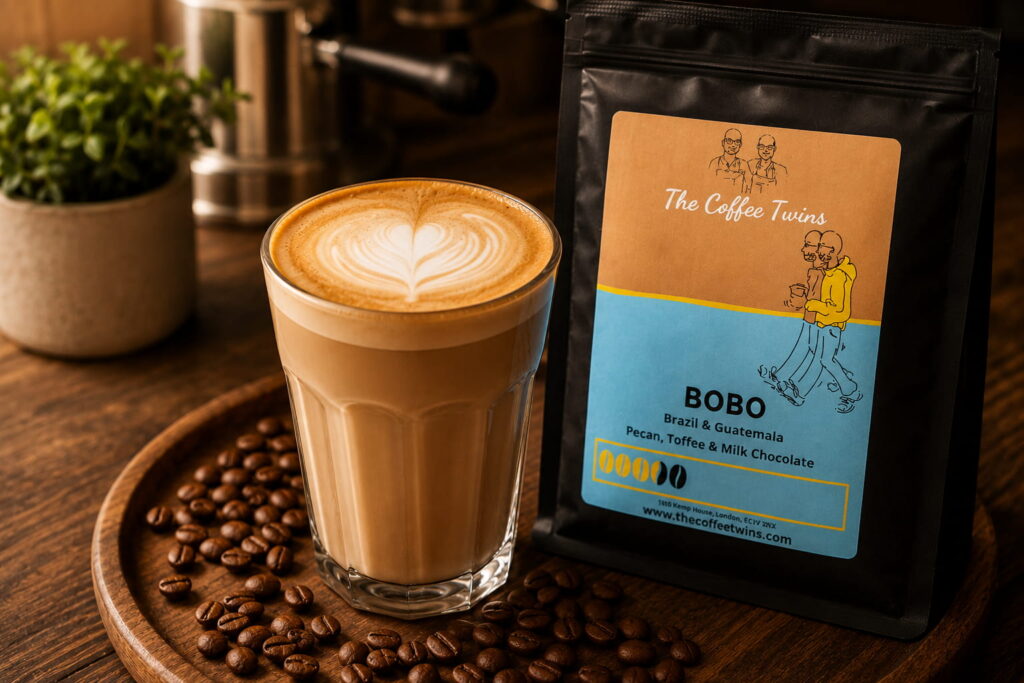 Creamy café-style latte with heart latte art served in a clear glass beside a bag of The Coffee Twins Bobo coffee beans on a wooden table with scattered coffee beans. The Bobo coffee bag features its official light blue label with notes of pecan, toffee and milk chocolate, creating a warm home coffee scene ideal for a guide on how to make a latte at home in the UK.