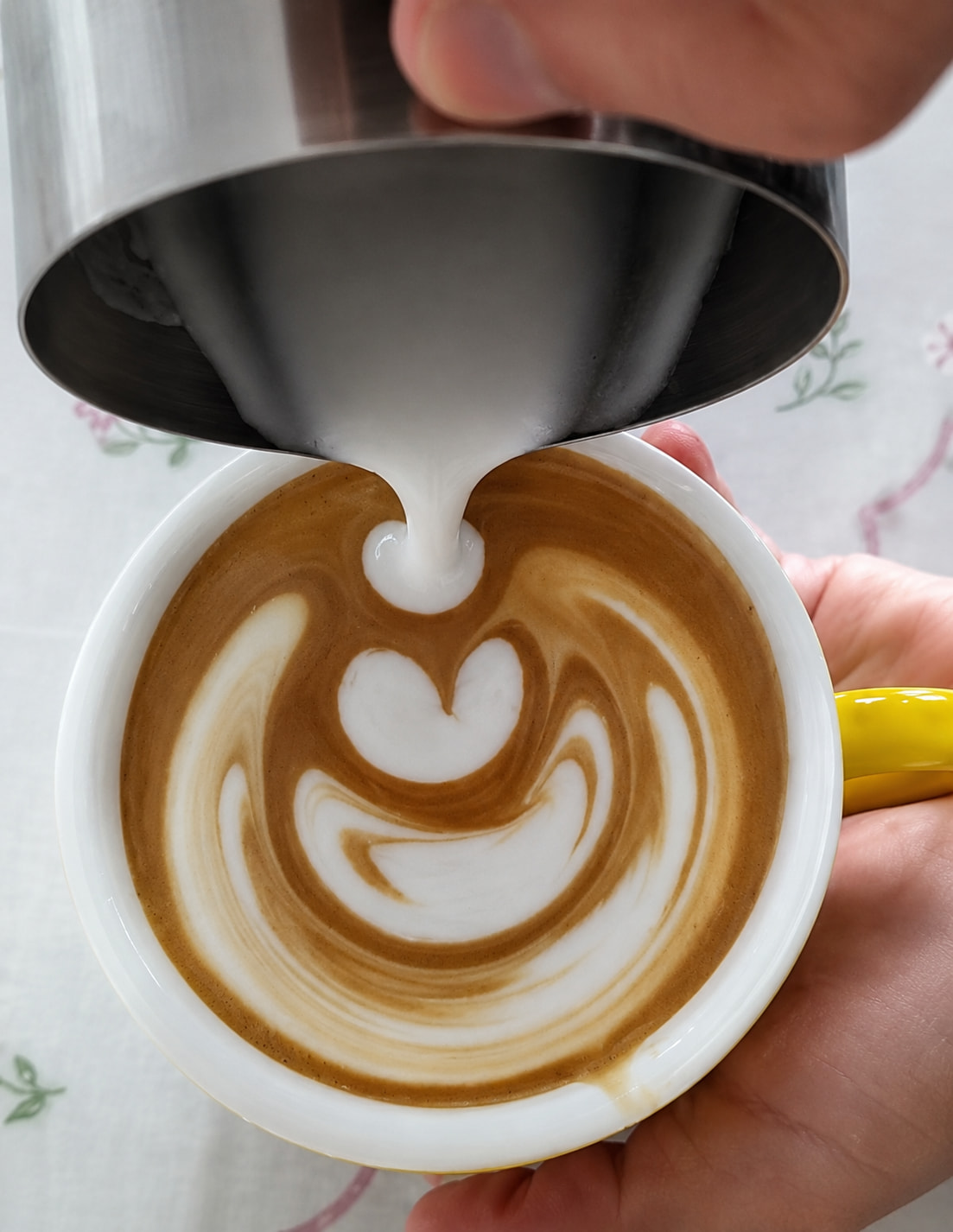 Close-up of a barista pouring steamed milk from a stainless steel pitcher into a white coffee cup to create tulip latte art. The image captures the process of making a latte with silky microfoam and espresso crema, demonstrating how to pour latte art at home using properly textured milk.
