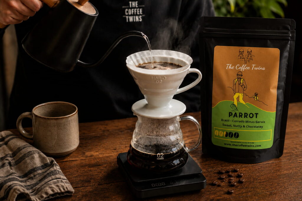 Hand pouring hot water over a Hario V60 pour-over coffee dripper beside The Coffee Twins Parrot speciality coffee beans, demonstrating the V60 coffee brewing method at home.
