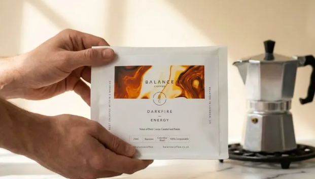 Hand holding Balance Coffee “Darkfire Energy” coffee pods in front of a moka pot on a kitchen stove, showcasing home coffee brewing.