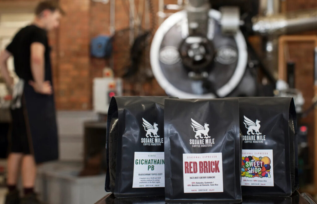 Three bags of Square Mile Coffee Roasters speciality coffee displayed in front of a coffee roaster machine, with a barista in the background, showcasing independent UK coffee roasting.