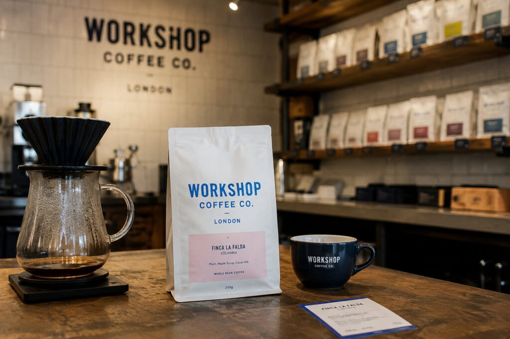 Workshop Coffee London bag on a café counter beside a pour-over dripper and cup, with shelves of speciality coffee bags in a modern UK coffee shop.