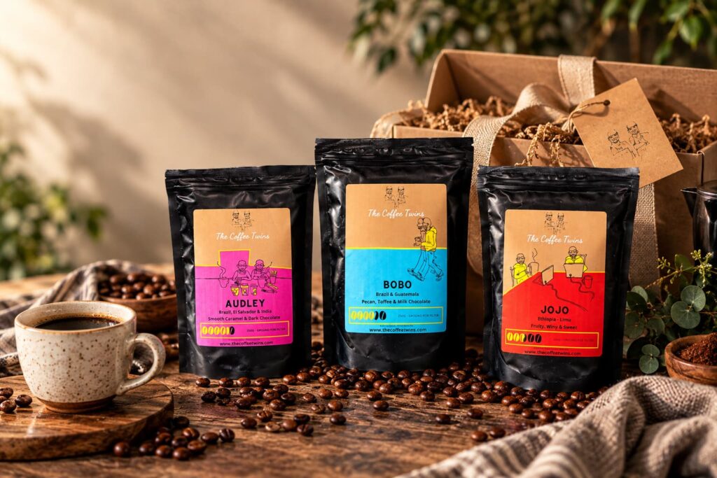 The Coffee Twins coffee tasting pack featuring Audley, Bobo and Jojo coffee bags displayed with roasted coffee beans, a cup of coffee and a gift box on a rustic wooden table.