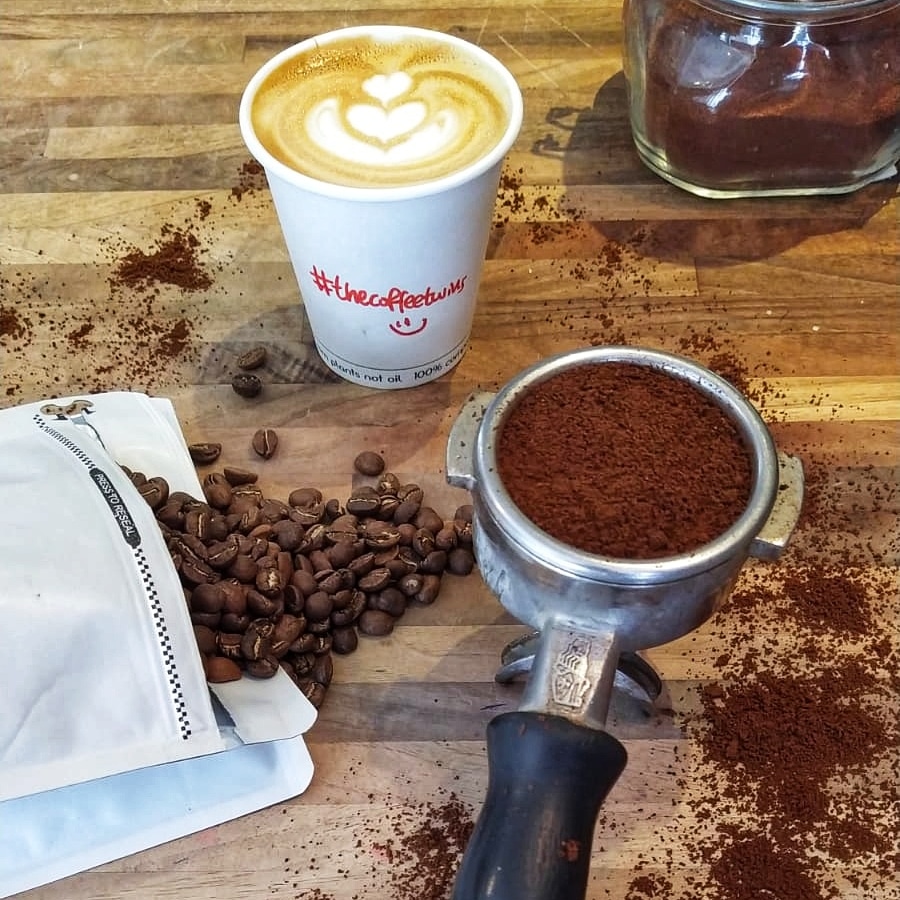 Coffee beans in a coffee bag being spilt onto the table, latte in a takeaway cup, ground coffee in a portafilter handle.