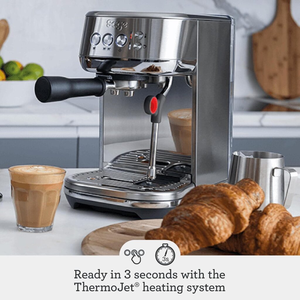 Stainless steel Sage Bambino espresso machine with freshly brewed coffee, milk pitcher and pastries on a kitchen counter.