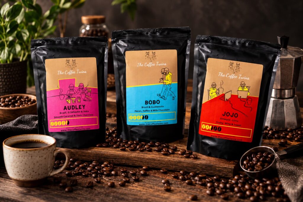 Three The Coffee Twins speciality coffee bags — Audley, Bobo and Jojo — displayed together with coffee beans and brewing equipment on a rustic wooden table.