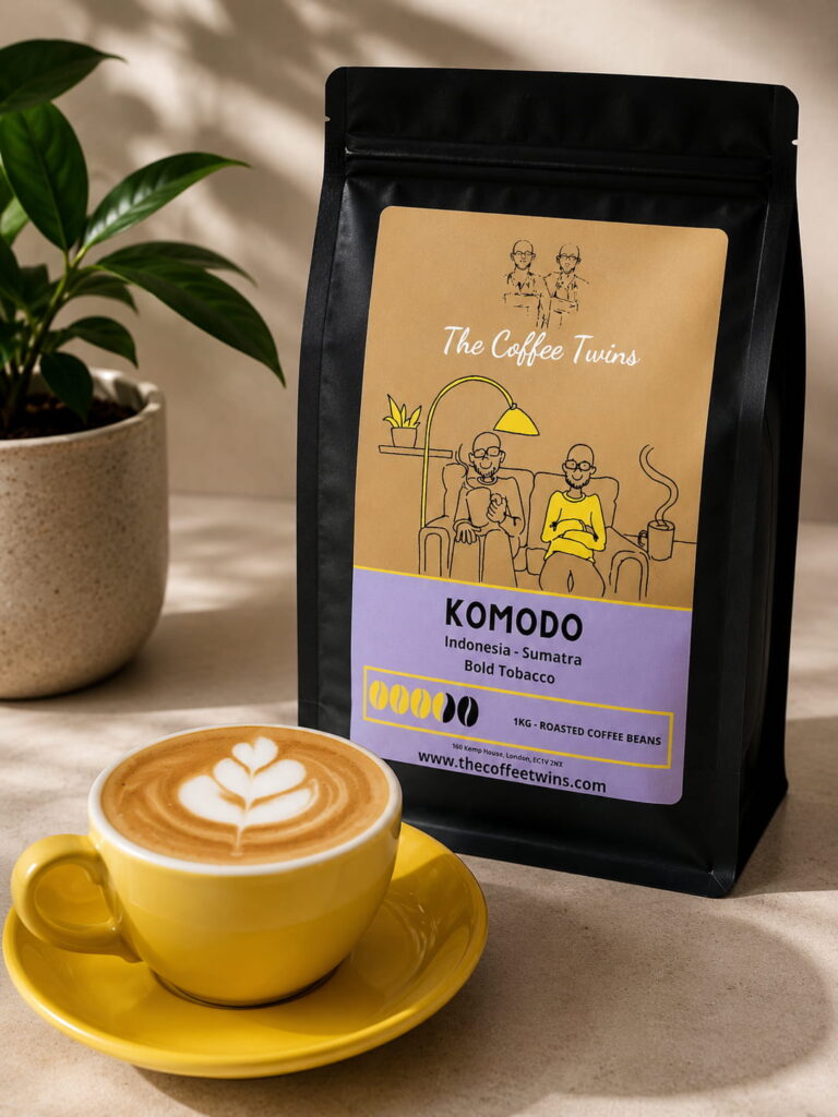 The Coffee Twins Komodo coffee bag with a freshly prepared latte in a yellow cup, showcasing Indonesian Sumatra roasted coffee beans in a warm, natural product photography setting.