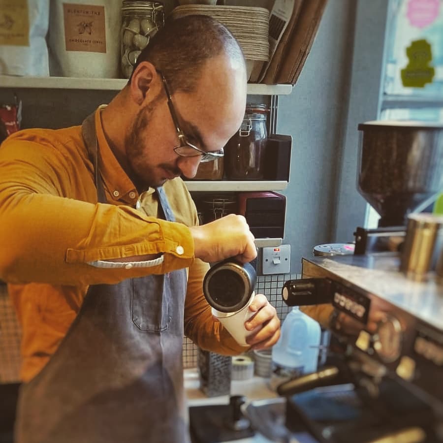 One of The Coffee Twins preparing speciality coffee in a café, carefully pouring steamed milk into a cup beside a professional espresso machine.